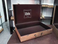 MALETA FLOCADA E MARROM RÚSTICO COM ALÇA E FECHO COM LOGO VAZADO NA TAMPA PARA IBIS HOTEIS