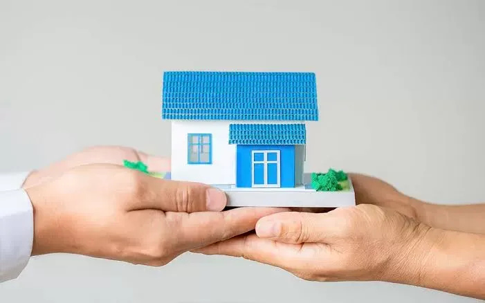Duas mãos apoiando juntas a miniatura de uma casa de telhado azul