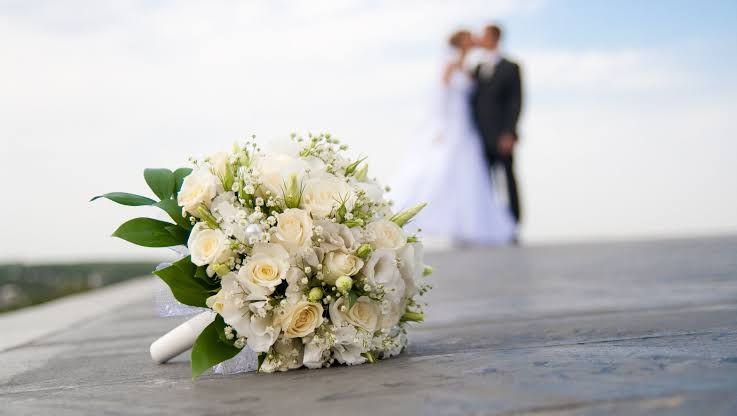 Buquê de rosas brancas em primeiro plano e, ao fundo desfocado, um casal de noivos
