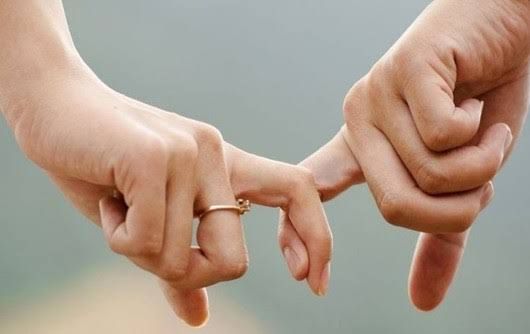 Mãos de duas pessoas com os dedos indicadores entrelaçados, uma delas usando anel