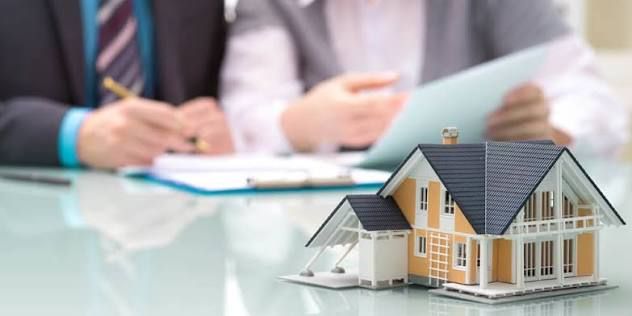 Miniatura de casa sobre a mesa e, ao fundo, duas pessoas de terno examinando documentos