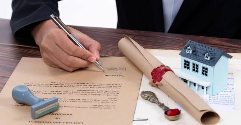Mão assinando um testamento sobre a mesa, ao lado de um pergaminho lacrado com selo de cera e de uma miniatura de casa
