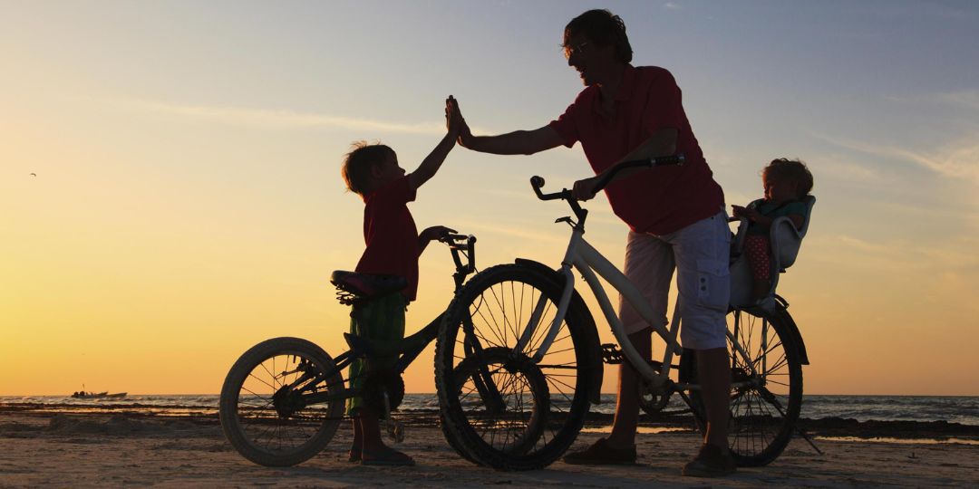 Adulto com duas crianças e bicicletas na praia ao pôr do sol, batendo as mãos com uma delas