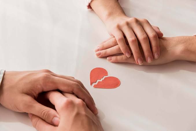 Duas pessoas sentadas à mesa com as mãos entrelaçadas e, entre elas, um coração de papel partido ao meio