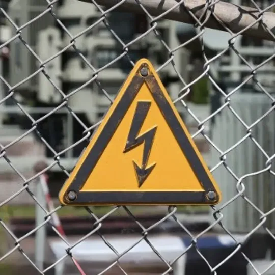 Placa de advertência de alta tensão fixada em grade de proteção em área industrial, sinalizando risco elétrico conforme normas da NR-10 sobre classificação de tensão.