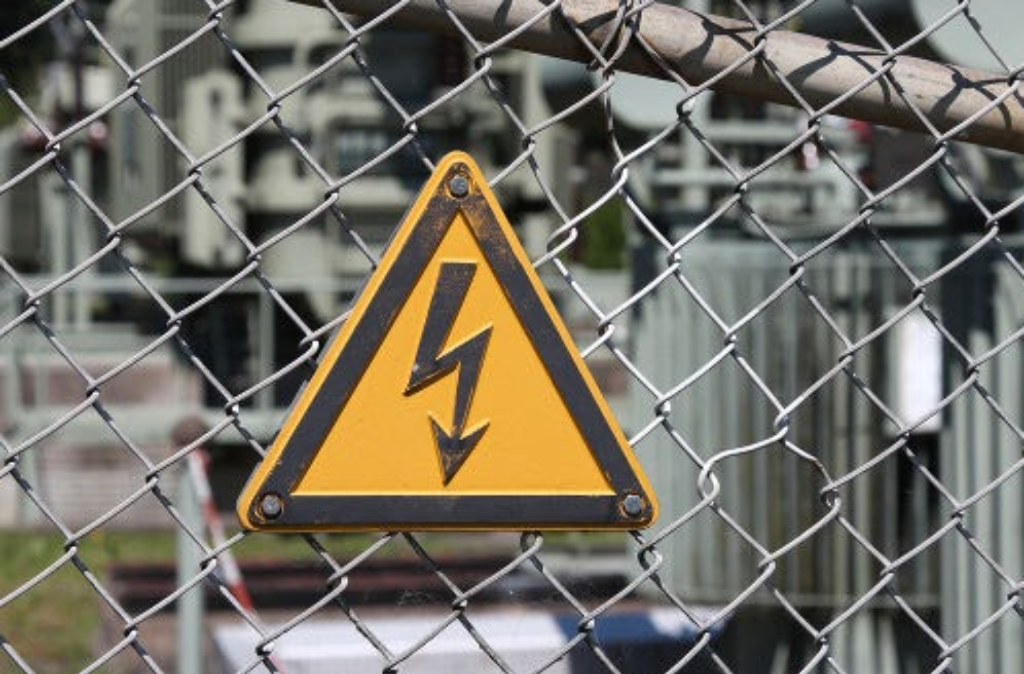Placa de advertência de alta tensão fixada em grade de proteção em área industrial, sinalizando risco elétrico conforme normas da NR-10 sobre classificação de tensão.