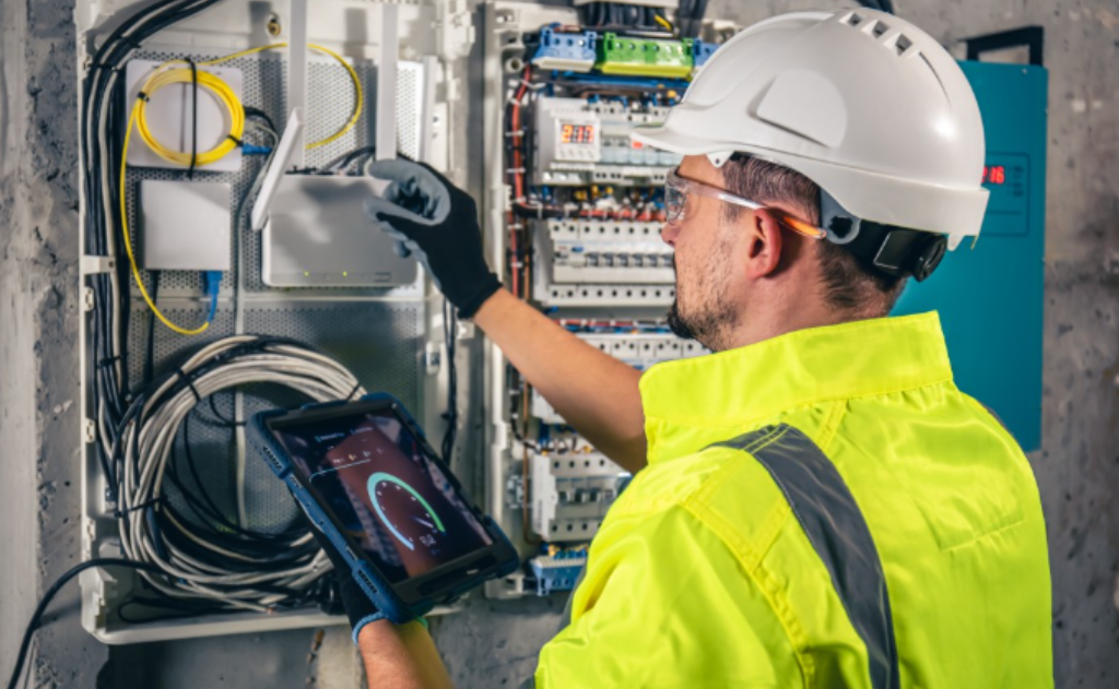 Técnico com EPI realizando manutenção preventiva em painel elétrico industrial, utilizando tablet para monitoramento de parâmetros em tempo real e garantir segurança e funcionamento contínuo do sistema.