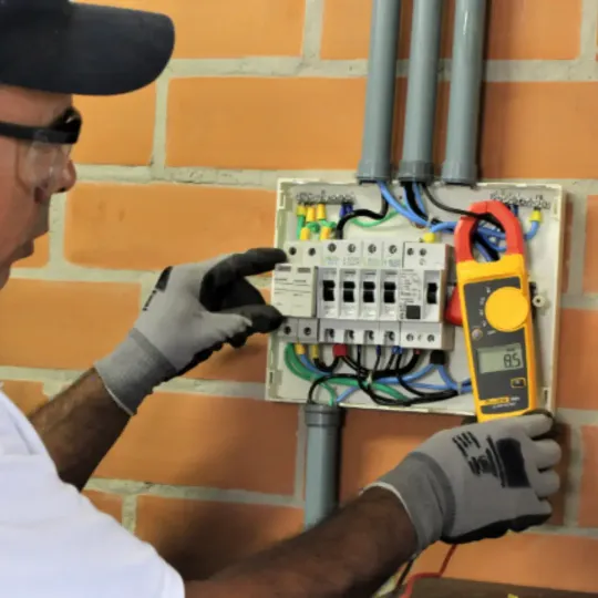 Eletricista utilizando alicate amperímetro para medir corrente em quadro de distribuição industrial, durante inspeção de queda de tensão em instalação elétrica. Imagem ilustrativa do processo de verificação para garantir eficiência e estabilidade elétrica em ambientes industriais.