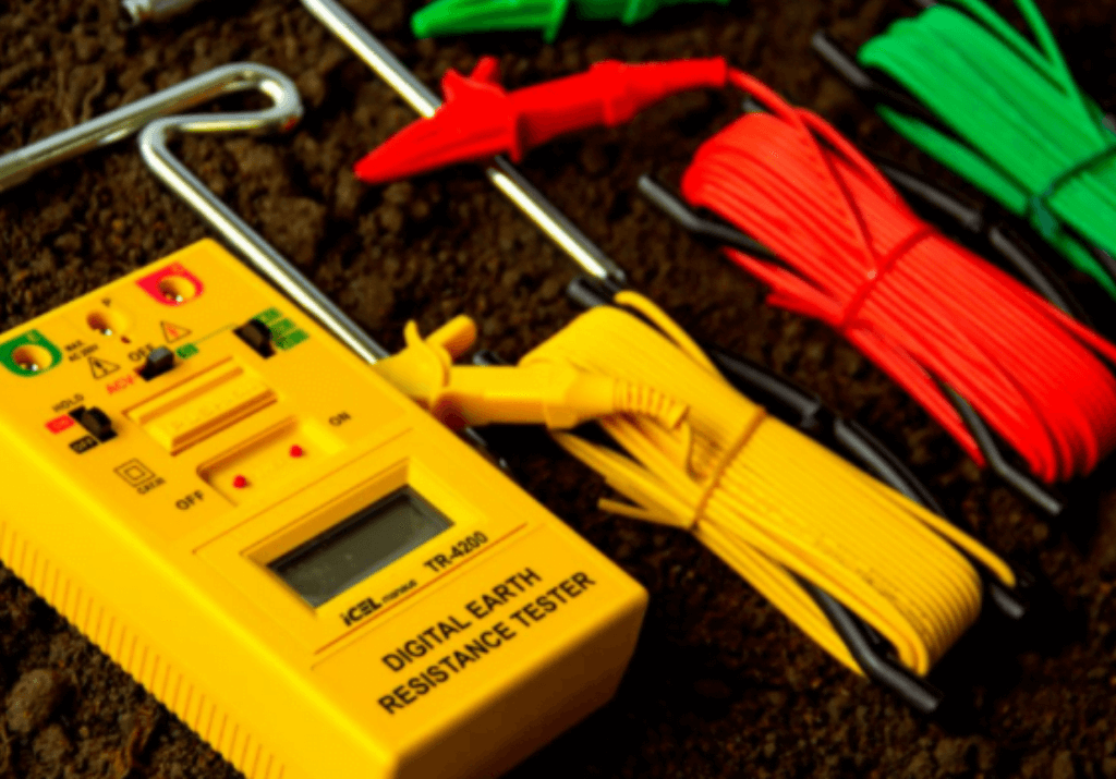 Medidor digital de resistência de aterramento com cabos e hastes de teste sobre o solo, utilizado para verificar a eficiência do sistema de aterramento em instalações elétricas.