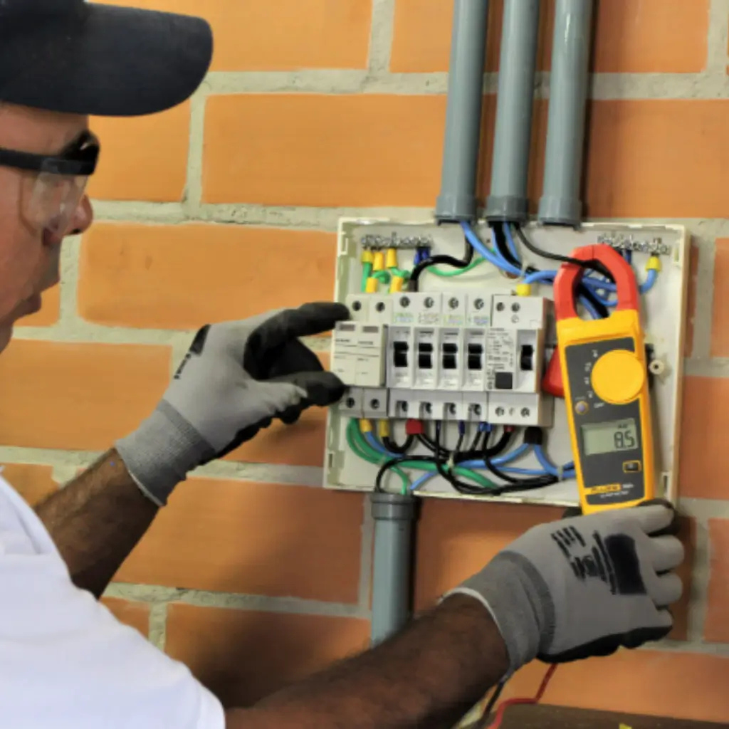 Eletricista utilizando alicate amperímetro para medir corrente em quadro de distribuição industrial, durante inspeção de queda de tensão em instalação elétrica. Imagem ilustrativa do processo de verificação para garantir eficiência e estabilidade elétrica em ambientes industriais.