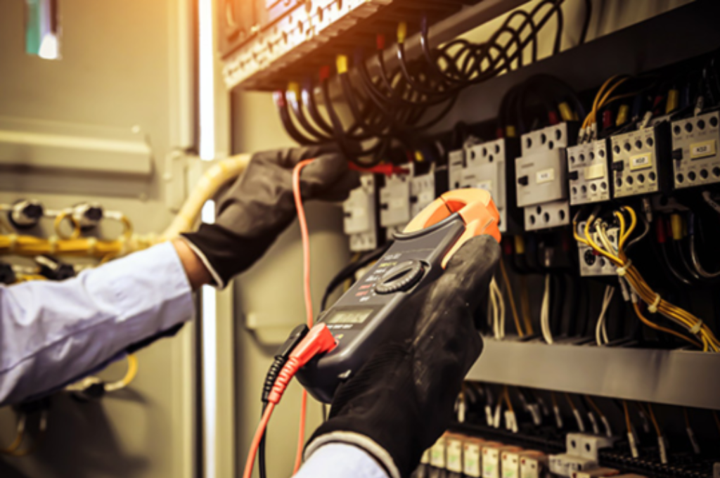 Técnico utilizando alicate amperímetro para medir corrente elétrica em painel de comando industrial, ilustrando procedimentos para evitar queda de tensão em instalações elétricas industriais.