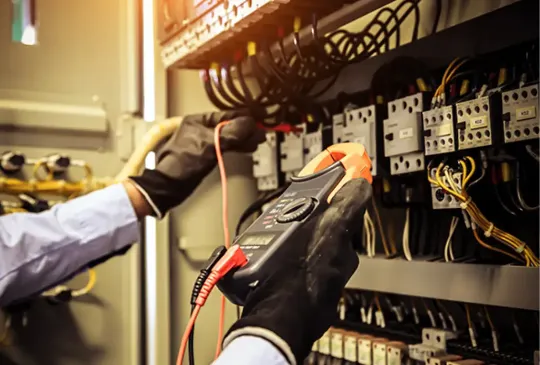 Técnico utilizando alicate amperímetro para medir corrente elétrica em painel de comando industrial, ilustrando procedimentos para evitar queda de tensão em instalações elétricas industriais.