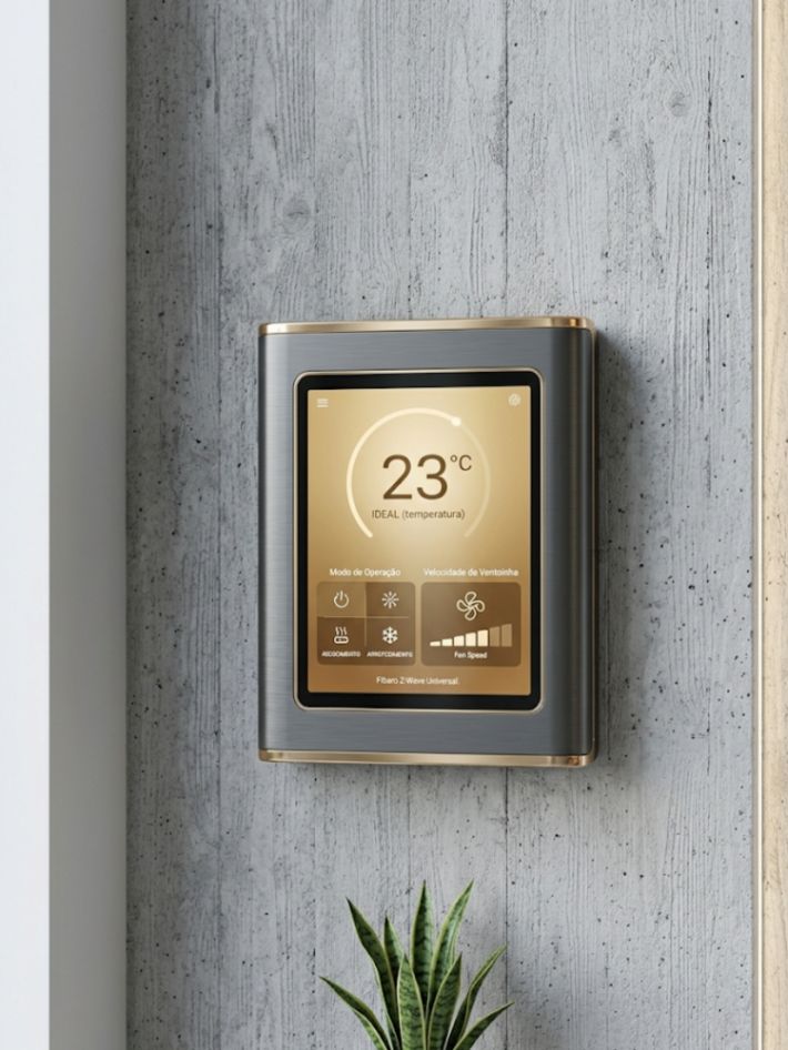 Fotografia vertical de termostato inteligente Fibaro com display tátil dourado mostrando temperatura ideal e controle de fan em parede minimalista de concreto aparente.
