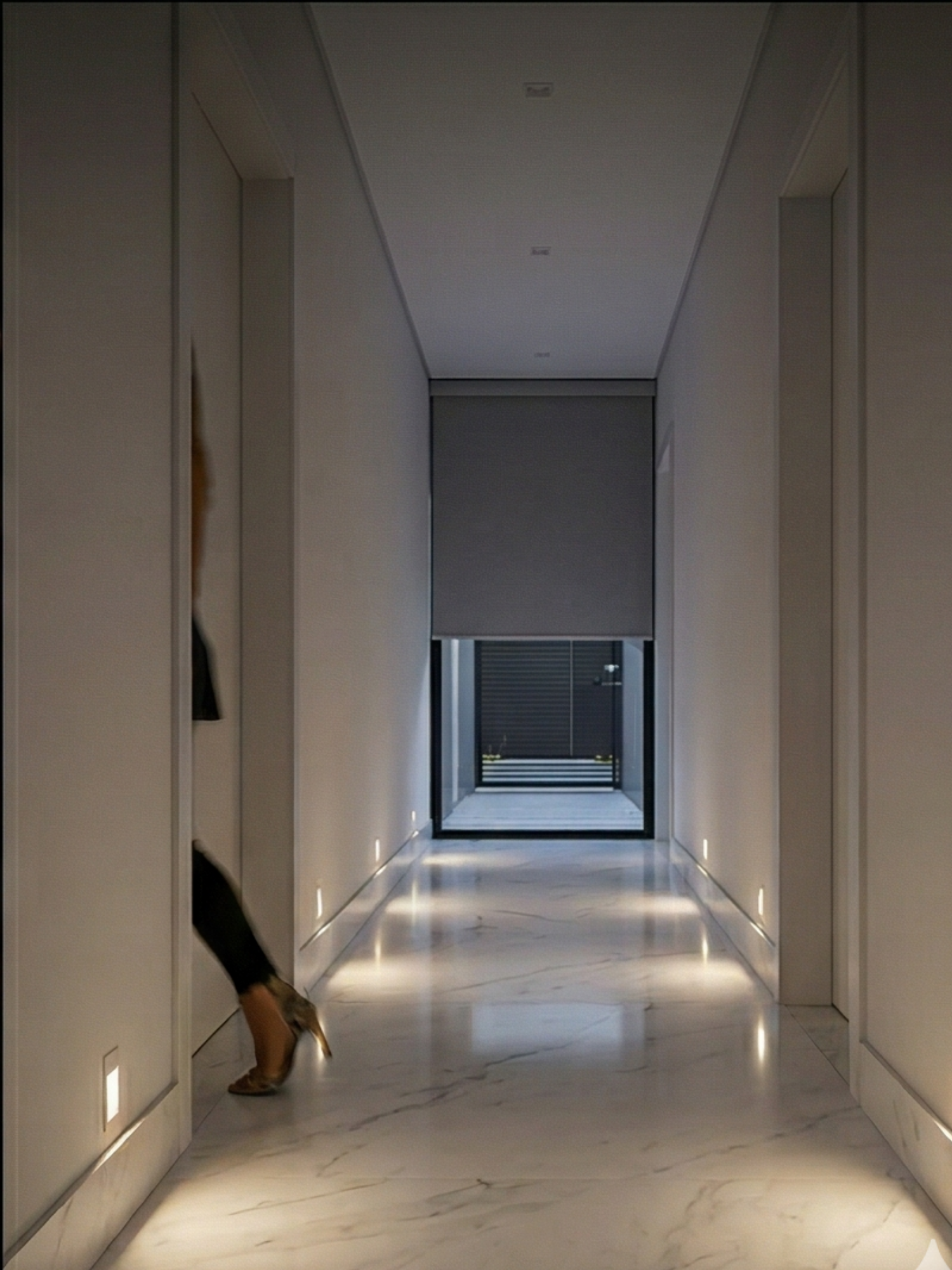 Close-up em um corredor minimalista com piso de mármore branco polido. Vemos os pés e pernas de uma pessoa caminhando, e balizadores de rodapé quentes se iluminam automaticamente à medida que ela passa. No final do corredor, uma persiana de rolo Nice motorizada descendo e um portão de garagem Nice são visíveis.